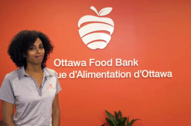 A woman stands in front of a red wall that reads, "Ottawa Food Bank" with its French translation below.