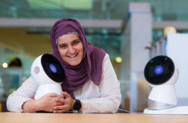 Sharifa Alghowinem, who wears a head scarf, leans on a table, hugging a Jibo personal robot, which looks like a sphere with a large eye.